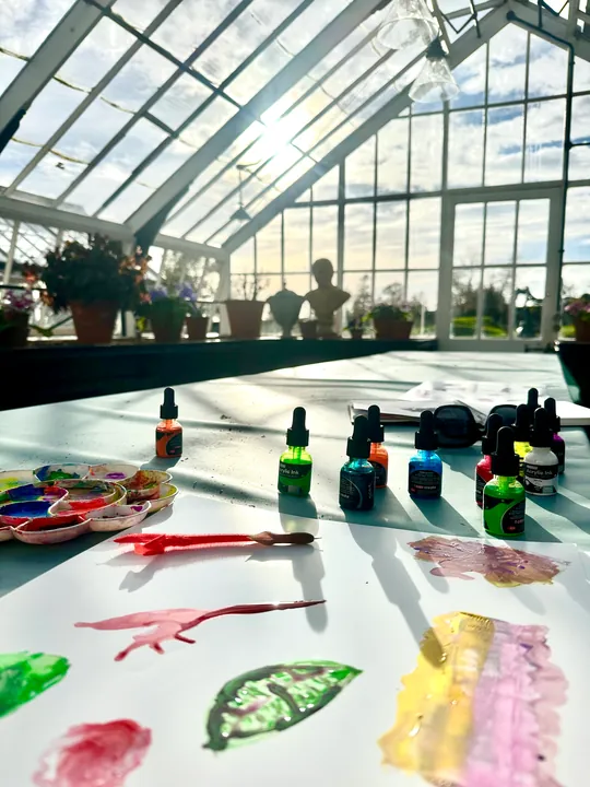 A table in a glasshouse with ink bottles, a palette and freshly painted marks