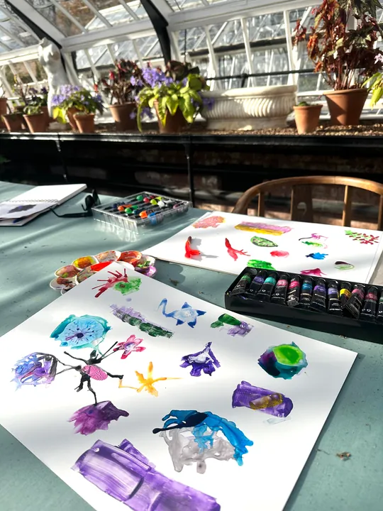 Sheets of painted marks laid out on a table in a glasshouse beside paint sets